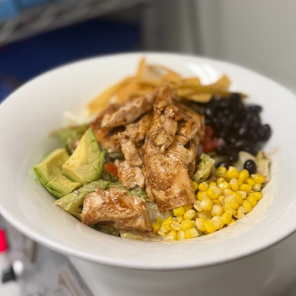A vibrant salad with grilled chicken, avocado, corn, black beans, and tortilla strips in a white bowl.