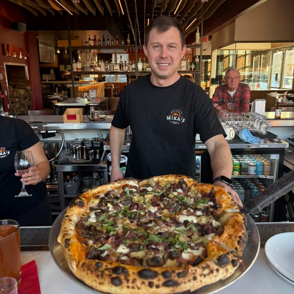 A Mikas Neapolitan Pizza chef proudly presents a large, freshly baked pizza with various toppings.