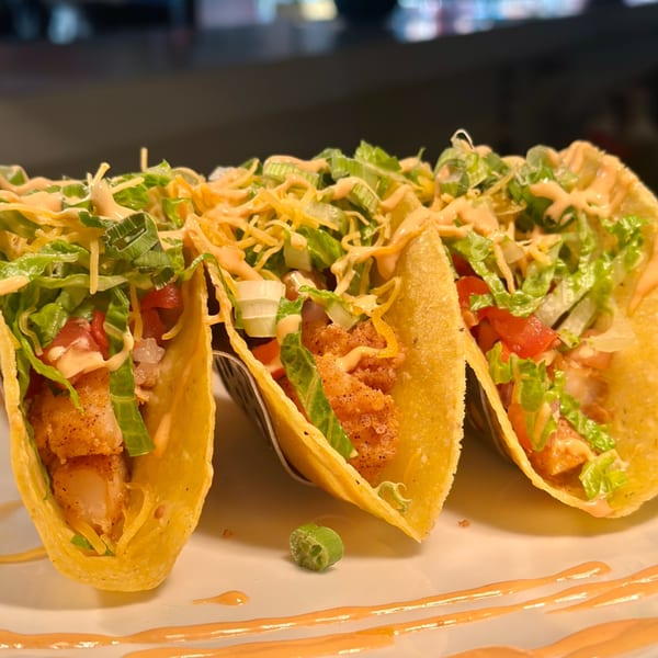 Three crispy shrimp tacos with lettuce, tomatoes, cheese, and a creamy sauce on a white plate.